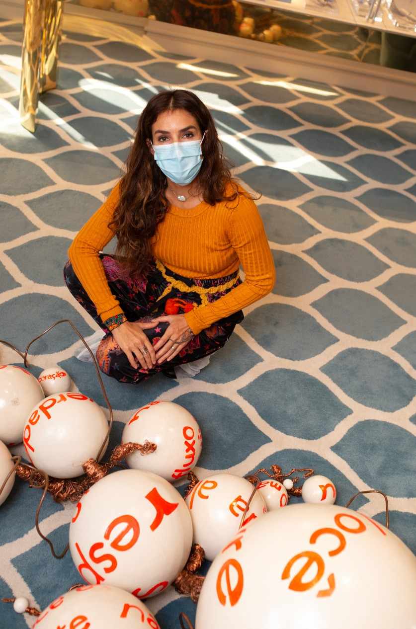 Graziella Pinto posa sentada atrás de sua obra feita de esferas