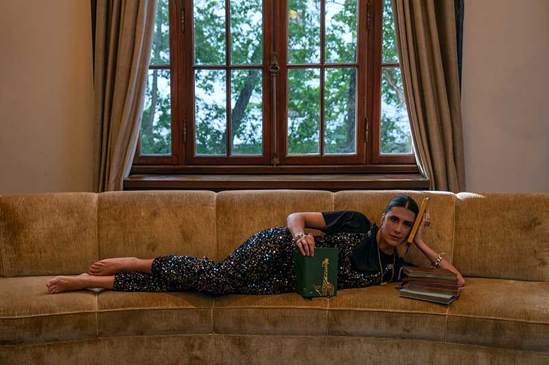 Maria Helena Pessôa de Queiroz, fundadora da MH Studios, posa deitada em um sofá segurando um livro