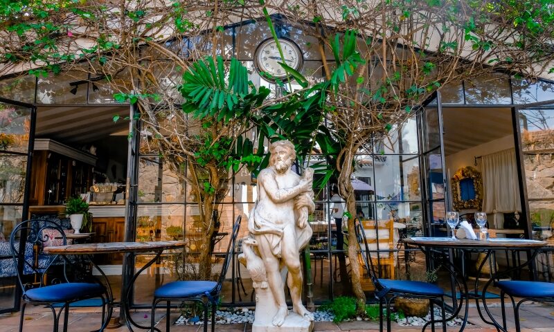Entrada do La Gare pelo pátio interno do hotel Vila da Santa, com o relógio garimpado pelos proprietários e a escultura em Carrara (divulgação)